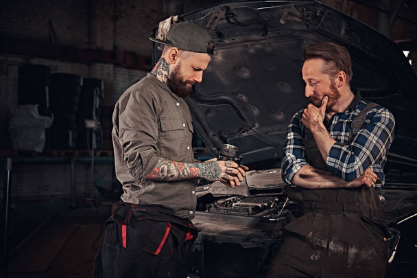 Два механика разговаривают во время ремонта сломанной машины в гараже
. - Фото, изображение