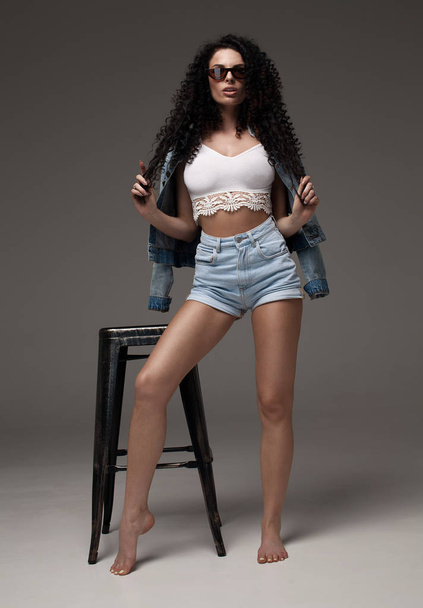 Sexy attractive brunette woman posing in fashionable jeans in studio. Fashion model posing. Long curly hair. - 写真・画像