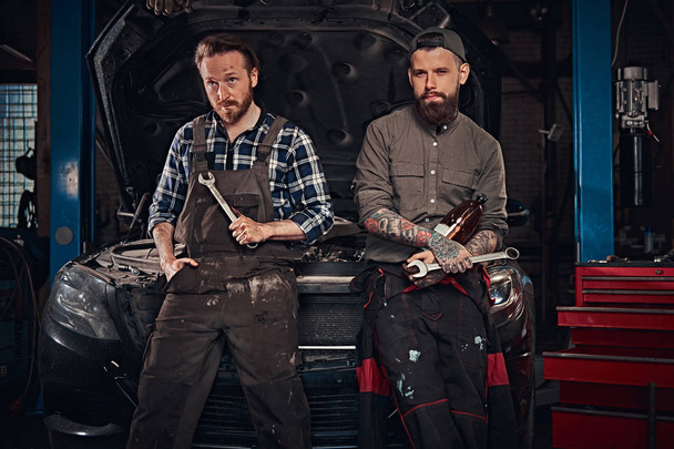 Two mechanic sit on a broken car, rest after a hard days work in a garage. - Fotoğraf, Görsel
