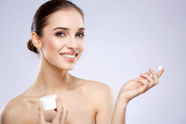young smiling natural woman applying cream on skin against grey background - Photo, Image