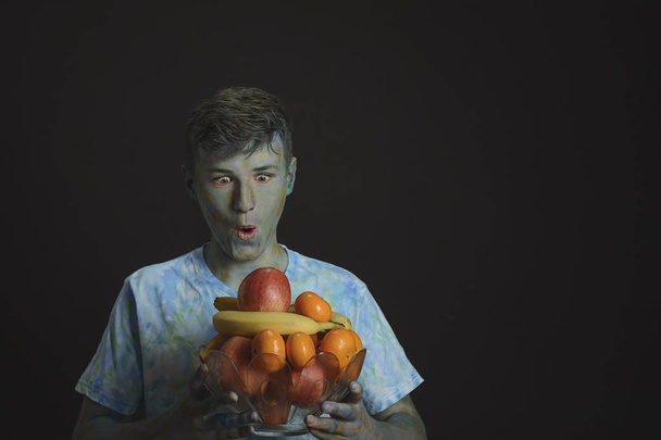 Painted multicolored paints young smiling guy holding a glass vase with fruit on a black background - Photo, Image