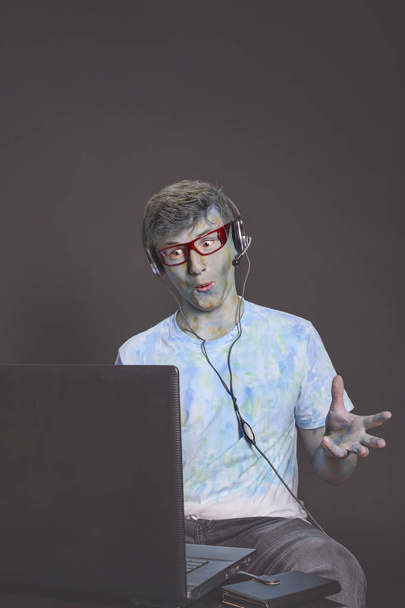 young and interesting guy, struck with multicolored paints in glasses and a headset, is talking animatedly through a computer - Foto, imagen