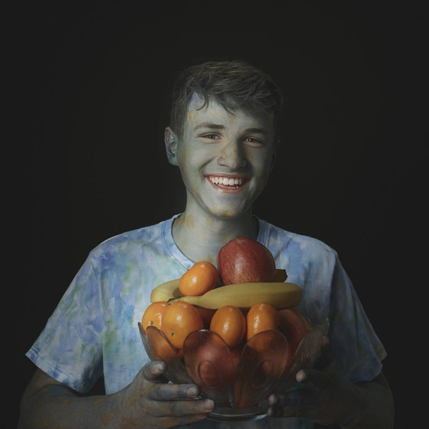 Peint multicolore peint jeune homme souriant tenant un vase en verre avec des fruits sur un fond noir
 - Photo, image
