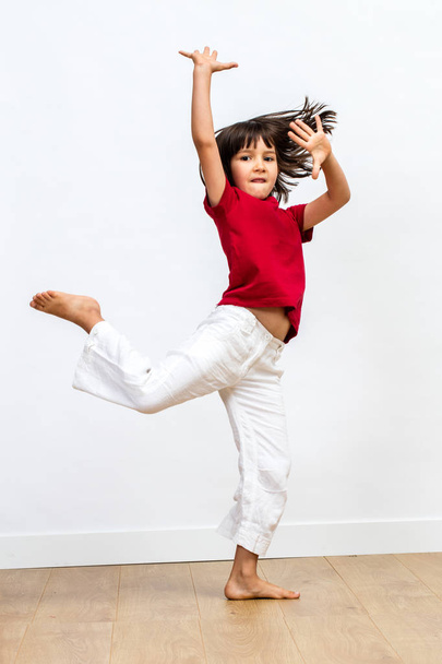 graciosa menina bonita com pés descalços dançando, mostrando movimento dinâmico e esportes alegres para expressar vitalidade infantil sobre piso de madeira, fundo branco
 - Foto, Imagem