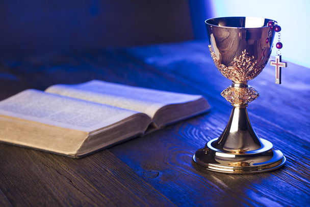 Roman Catholic Church theme. Holy Bible, rosary and golden chalice on brown wooden table. - Photo, Image