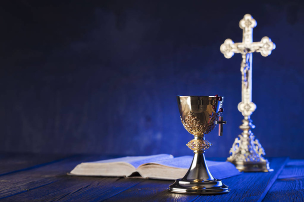 Тема Римско-католической церкви. Крест, Священная Библия, четки и золотая чаша на коричневом деревянном столе
. - Фото, изображение