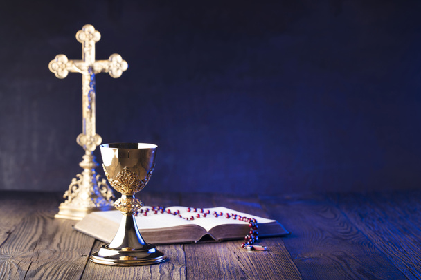 Roman Catholic Church theme. The Cross, Holy Bible, rosary and golden chalice on brown wooden table. - Photo, Image