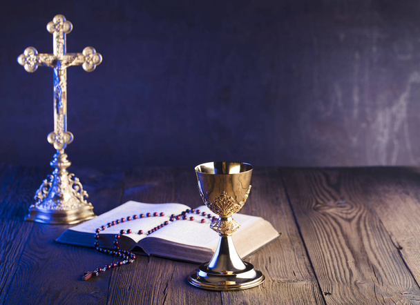 Thema der römisch-katholischen Kirche. Kreuz, Bibel, Rosenkranz und goldener Kelch auf braunem Holztisch. - Foto, Bild