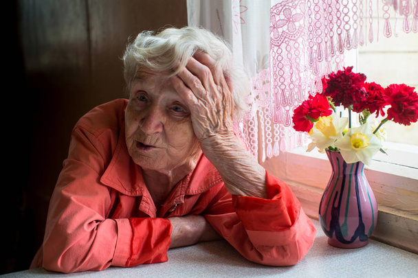 Ηλικιωμένη γυναίκα φαίνεται δυστυχώς να κάθεται κοντά στο παράθυρο. - Φωτογραφία, εικόνα