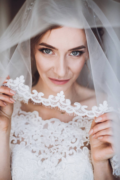 Beautiful blonde bride in elegant white lace wedding dress and veil holding sexy garter portrait, face closeup of gorgeous bride in hotel room - Photo, Image