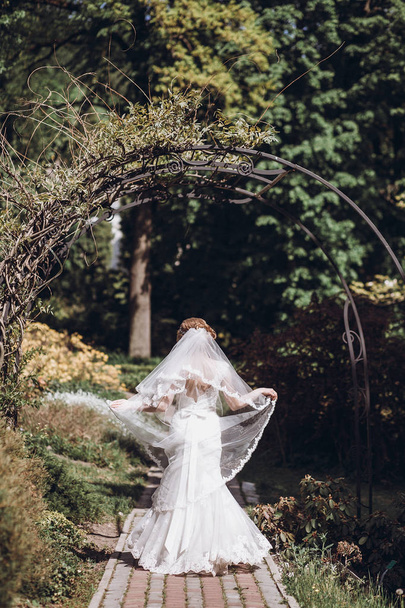 πανέμορφη νύφη με πέπλο θέτοντας και τη διασκέδαση στο πάρκο ηλιόλουστη άνοιξη. ευτυχισμένη γυναίκα στέκεται μέσα σε καταπράσινο κήπο. στιγμές ευτυχισμένο γάμο. δραπέτευση έννοια - Φωτογραφία, εικόνα