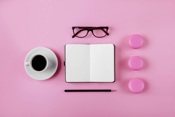 Coffee, macaron, clean notebook, eyeglasses and flower on pink table from above. Female working desk. Flat lay style. - Foto, afbeelding