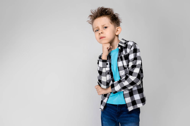 Un chico guapo con camisa a cuadros, camisa azul y pantalones vaqueros está parado sobre un fondo gris. El niño sostiene su barbilla con su mano
 - Foto, Imagen