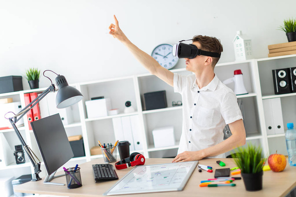 A young man in virtual reality glasses stands near the table and pulls his arm up. A young man thumbs a virtual page. - Photo, image