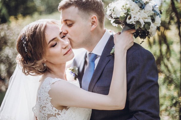 beautiful gorgeous bride and groom kissing in sunny spring park and enjoying day. happy wedding couple relaxing in green garden. happy marriage moments. elopement concept - Photo, Image