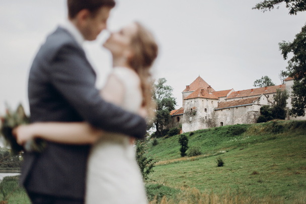όμορφη πανέμορφη νύφη και ο γαμπρός το περπάτημα στο ηλιόλουστο πάρκο και φιλιά. ευτυχισμένος γάμος ζευγάρι αγκαλιάζει μέσα σε καταπράσινο κήπο στο παλιό κάστρο. επικεντρωθούν στην οικοδόμηση. δραπέτευση έννοια - Φωτογραφία, εικόνα