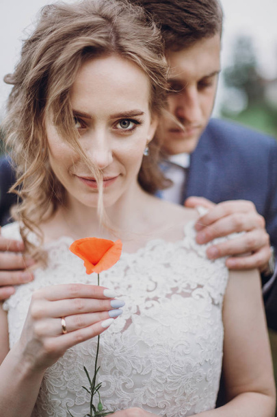 Bräutigam umarmt schöne wunderschöne Braut mit Mohnblume. glückliches, sinnliches Hochzeitspaar, das sich verliebt auf einem alten Schloss umarmt. glückliche romantische Momente. Elopement-Konzept. Raum für Text - Foto, Bild