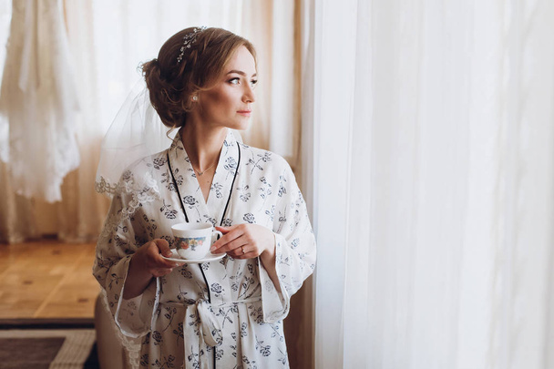 Espléndida novia en elegante túnica bebiendo café en hotel de lujo cara a cara primer plano de expresión de ensueño, mañana antes de la preparación de la boda, reflejo de la ventana
 - Foto, Imagen