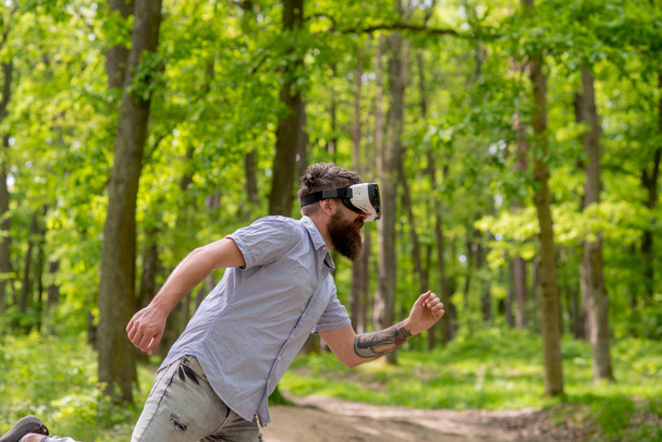 Hombre barbudo que tiene experiencia en 3D con auriculares VR. Vista lateral hipster barbudo en el bosque. Hombre con barba y bigote de moda corriendo en realidad virtual, artilugios modernos, concepto de mundo digital
. - Foto, Imagen
