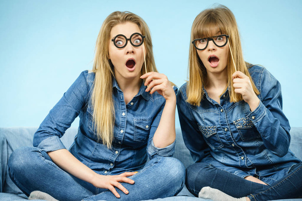 Two happy women holding fake eyeglasses on stick having shocked face expression wearing jeans shirts. Photo and carnival funny accessories concept. - Foto, Imagem