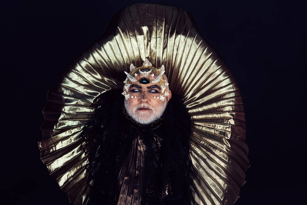 Ancient king of fairy tale land sitting on golden throne. Old bearded blind man with three eyes and thorns on his head isolated on black background. Wizard with demonic appereance predicting future. - Foto, imagen