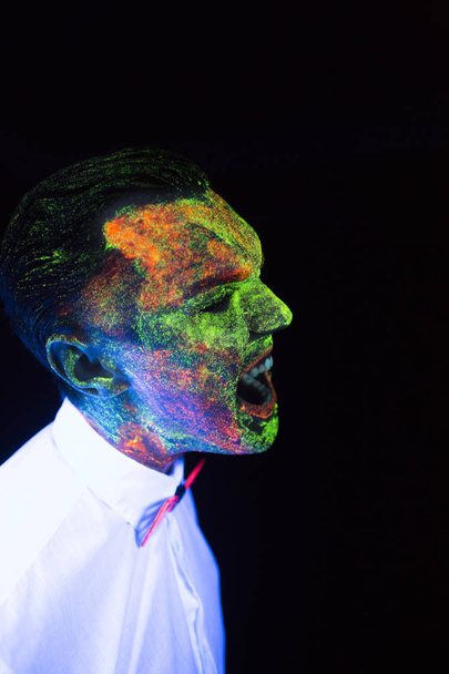 concept of wedding photosession in neon lighting in vogue style groom young guy screams ana roars glows in the dark posing in white wedding suit with a bow tie under the UV rays on a dark background - Photo, Image