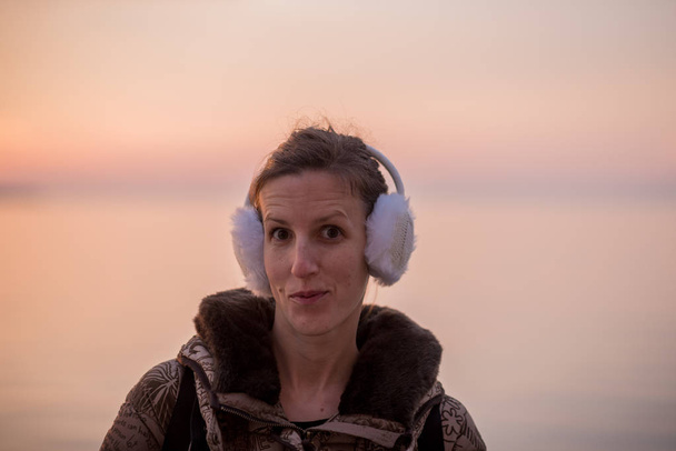 Jeune femme portant des chauffe-oreilles moelleux lors d'une soirée froide debout regardant la caméra contre un ciel couchant
. - Photo, image