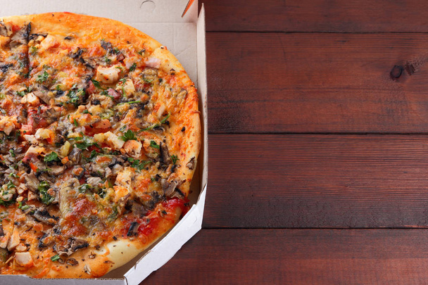 Pizza-ban egy doboz, egy fából készült háttér. Olasz pizza, csirke, gomba, zöldek, sajt és mártással. Ünnepi étkezés egy karton dobozban - Fotó, kép
