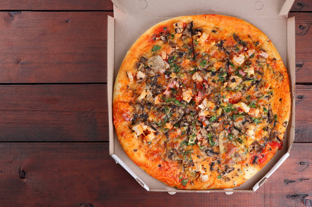Pizza in a box on a wooden background. Italian pizza with chicken, mushrooms, greens, cheese and sauce. Festive meal in a cardboard box - Photo, image