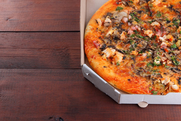 Pizza in a box on a wooden background. Italian pizza with chicken, mushrooms, greens, cheese and sauce. Festive meal in a cardboard box - 写真・画像