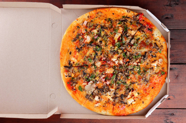 Pizza in a box on a wooden background. Italian pizza with chicken, mushrooms, greens, cheese and sauce. Festive meal in a cardboard box - 写真・画像