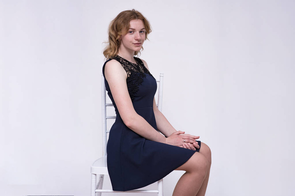 portrait of a young beautiful blonde girl on a white background sitting on a chair. She sits right in front of the camera and looks happy - Valokuva, kuva