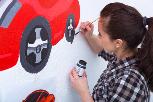 Young woman painter and mother boy draws for a child on a white wall a beautiful red car in a light children's room - Photo, Image