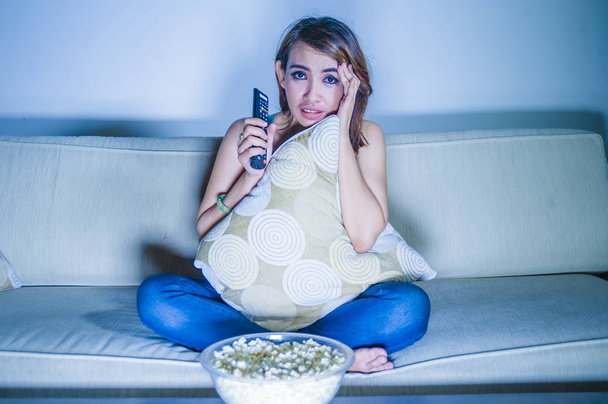 young beautiful sad latin woman watching drama romantic movie eating popcorn sitting at home sofa couch late night in sadness face expression holding pillow feeling emotional - Photo, Image