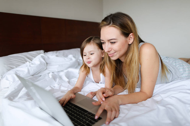 Nuori eurooppalainen nainen käyttää kannettavaa tietokonetta, jolla on pieni tytär ja joka makaa sängyssä
. - Valokuva, kuva