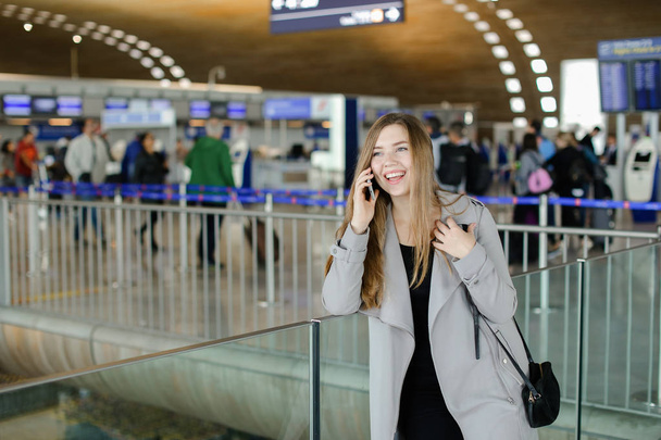 Красива жінка, розмовляючи за допомогою смартфона в залі аеропорту, одягнена в сіре пальто і сумку
. - Фото, зображення
