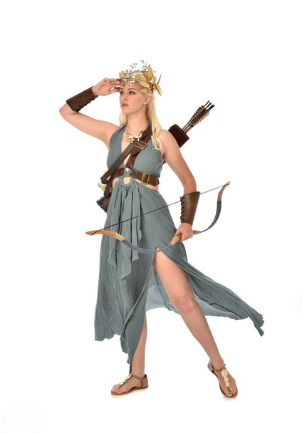 full length portrait of pretty blonde lady wearing fantasy toga gown,  and holding a bow and arrow. standing pose on white background. - Фото, изображение