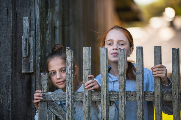 Due sorelle ragazze guardano fuori da dietro una recinzione di legno villaggio
. - Foto, immagini