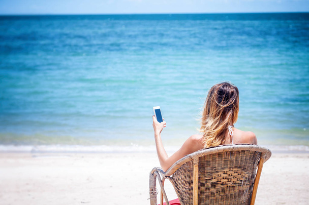 A girl in a white bikini with a mobile phone in hands on the shore of a tropical beach, a connection during a vacation and travel - Photo, Image