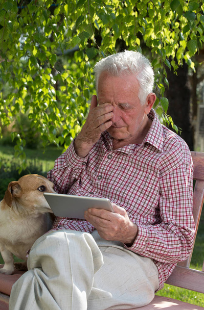 Smutny starszy mężczyzna z psem, siedząc na ławce w ogrodzie, patrząc na tablet i płacz - Zdjęcie, obraz