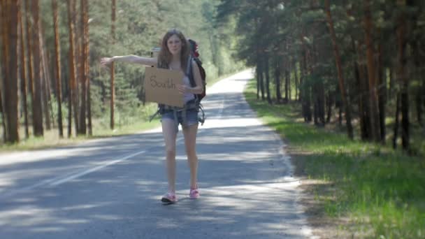 Młoda kobieta piękne autostopem stojący na drodze z plecakiem na stół z napisem południe - Materiał filmowy, wideo
