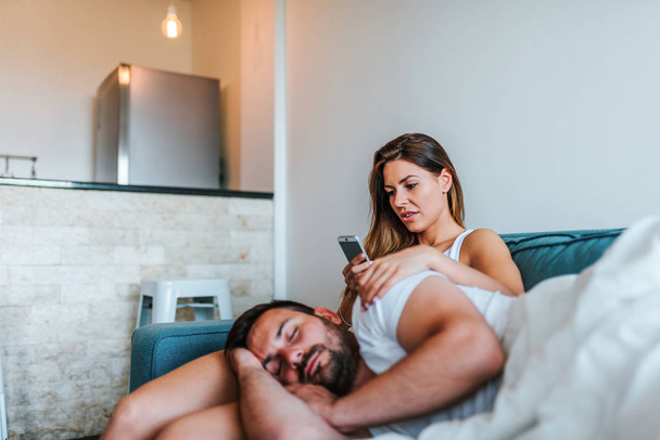 Jelous young woman cheking boyfriend's phone while he sleeps. - Zdjęcie, obraz