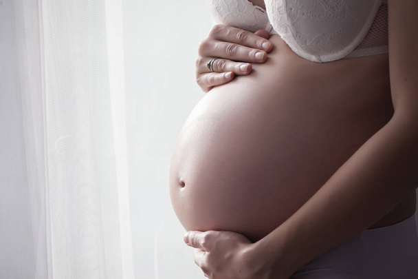 Bauch einer schwangeren Frau in weißer Unterwäsche, die neben den Fenstervorhängen steht - Foto, Bild