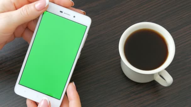 Ženské ruce s krásnou francouzskou manikúru si bílý smartphone s Green Screen poblíž bílý šálek kávy. Pomocí Smartphone, držení Smartphone s Green Screen na stylový černý dřevěný stůl - Záběry, video
