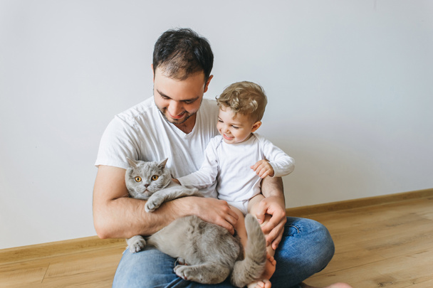 家でボディー スーツに幼い息子と手で灰色のブリティッシュショートヘアの猫を抱えて笑顔の父 - 写真・画像