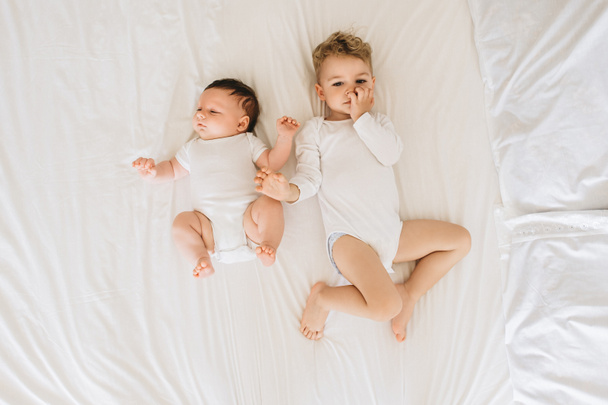 overhead view of cute little brothers in white bodysuits lying on bed together at home - Photo, Image