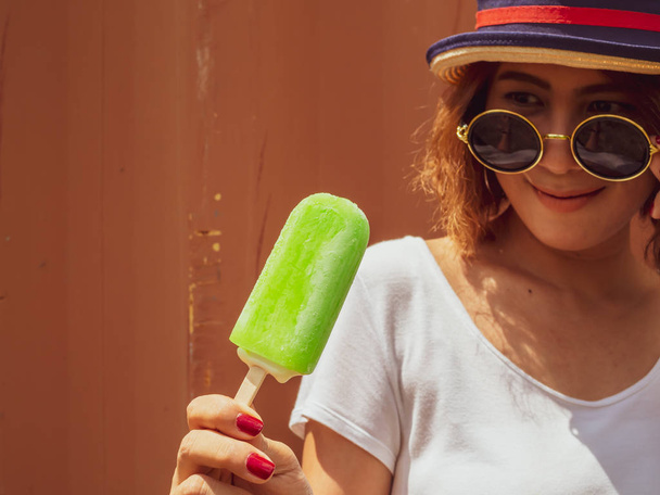 Beautiful Woman Short Hair Wearing White Shirt, Hat and Round Sunglasses with Happy Smiling Holding Green Frozen Popsicle Ice Pop on Summer Time with Red Container Background - Photo, Image