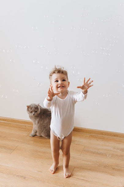 niño sonriente en traje de cuerpo blanco atrapando burbujas de jabón con gato gris cerca en casa
 - Foto, imagen