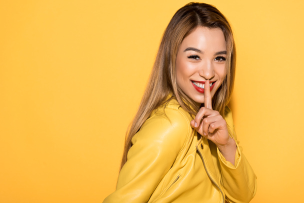 smiling asian female model closing mouth by finger isolated on yellow background - Foto, imagen