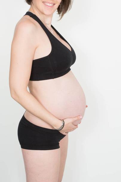 Close up on unidentifiable single pretty pregnant woman in sport clothes holding belly in front of gray background - Foto, imagen
